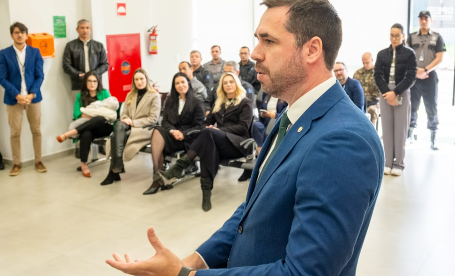 O Defensor Público-Geral é retratado de um ângulo diferente, discursando para um público maior. Ele está em primeiro plano e de perfil, com as mãos abertas num gesto expressivo. Ao fundo, uma plateia de aproximadamente quinze pessoas, composta por homens e mulheres de diversas idades, está sentada e acompanha o discurso com atenção.