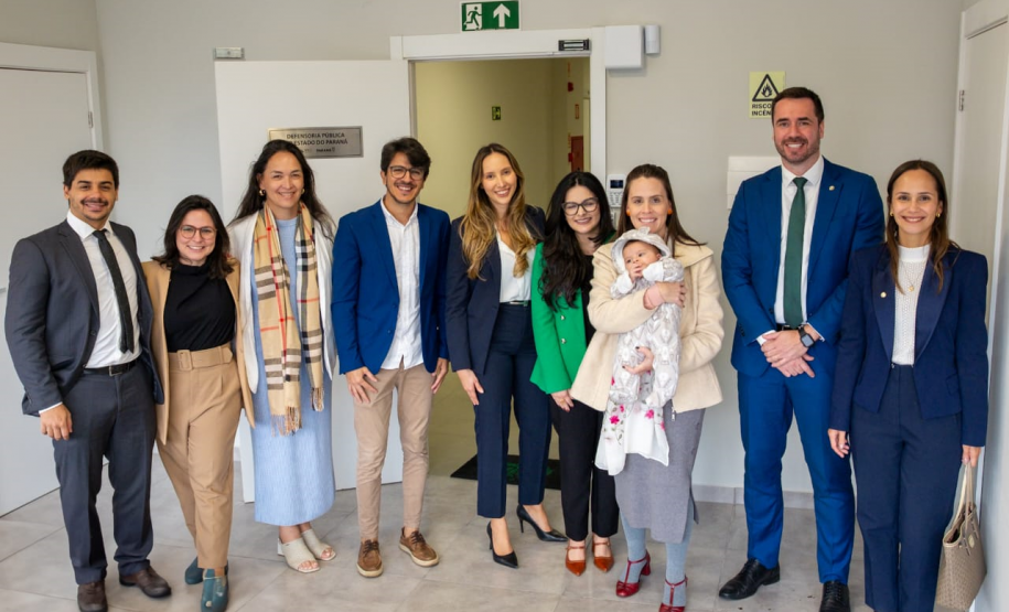Esta é uma foto de confraternização, que reúne um grupo posando para a câmera. Estão presentes a Ouvidora-Geral, defensores da sede de Foz, o Defensor Público-Geral e a Primeira e Segunda Subdefensoras-Gerais. São dez pessoas no total, além de um bebê no colo de uma das participantes. Todos sorriem em um corredor do novo prédio, onde uma placa na parede identifica o local: "DEFENSORIA PÚBLICA DO ESTADO DO PARANÁ".