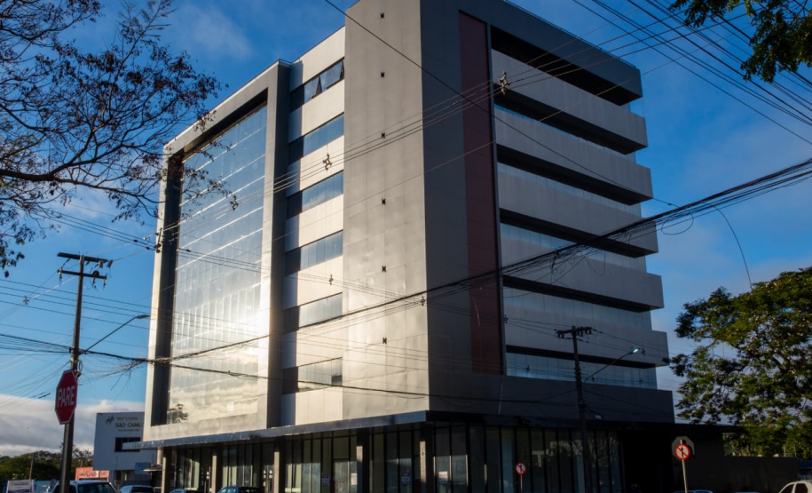 Esta foto exibe edifício da sede atual da DPE-PR em Foz do Iguaçu. Trata-se de um prédio grande e moderno de múltiplos andares. O céu está azul e ensolarado. A fachada do prédio é uma combinação de vidro espelhado, que reflete o céu, e painéis de cor cinza-escuro. A arquitetura tem linhas retas e um design contemporâneo.