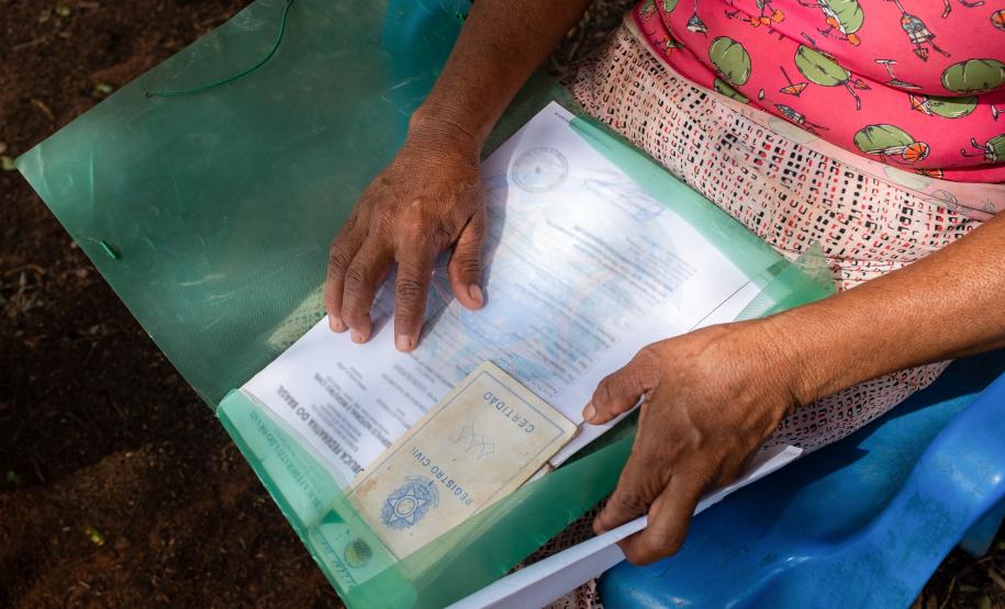Imagem mostra em detalhe os braços de uma mulher segurando no colo uma pasta com seus documentos. Ela está sentada numa cadeira de plástico e ao fundo se vê o chão de barro.