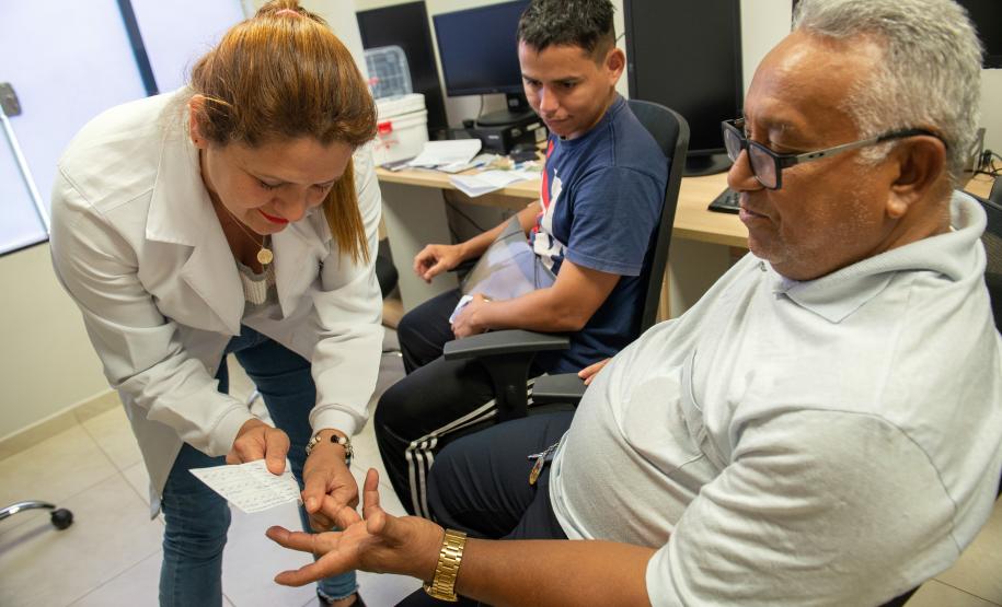 Imagem mostra um pai já idoso colhendo amostra de DNA com a enfermeira enquanto o filho já adulto observa a cena muito emocionado durante Mutirão Meu Pai Tem Nome da DPE-PR.