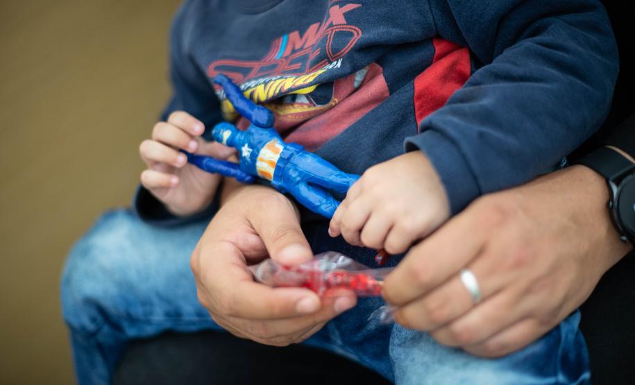 Imagem mostra em close uma criança sentada no colo do pai segurando um boneco de super herói enquanto a aguarda atendimento no Mutirão Meu Pai Tem Nome.