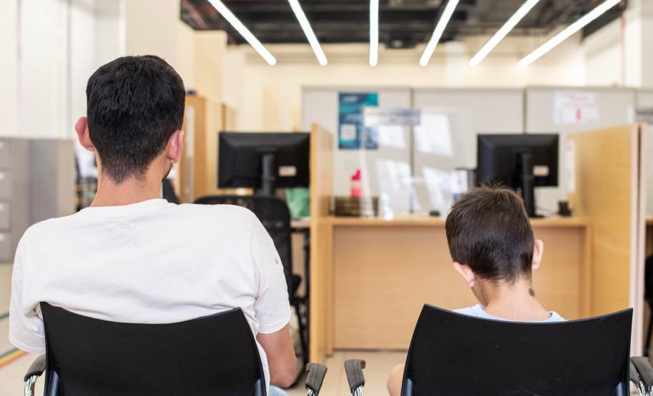 Imagem mostra pai e filho sentados de costas aguardando atendimento da DPE-PR no mutirão Meu Pai Tem Nome.