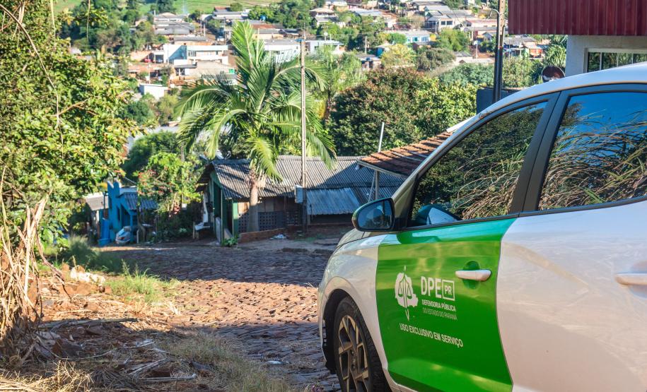 Imagem mostra carro da DPE-PR em rua calçada com pedras e ao fundo casas em meio à plantações.