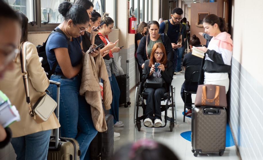 Imagem mostra uma candidata cadeirante chegando para uma das fases do V Concurso para Defensora e Defensor Público do Estado do Paraná. No primeiro plano fora de foco é possível ver vários candidatos aguardando para entrar na sala enquanto ela chega.