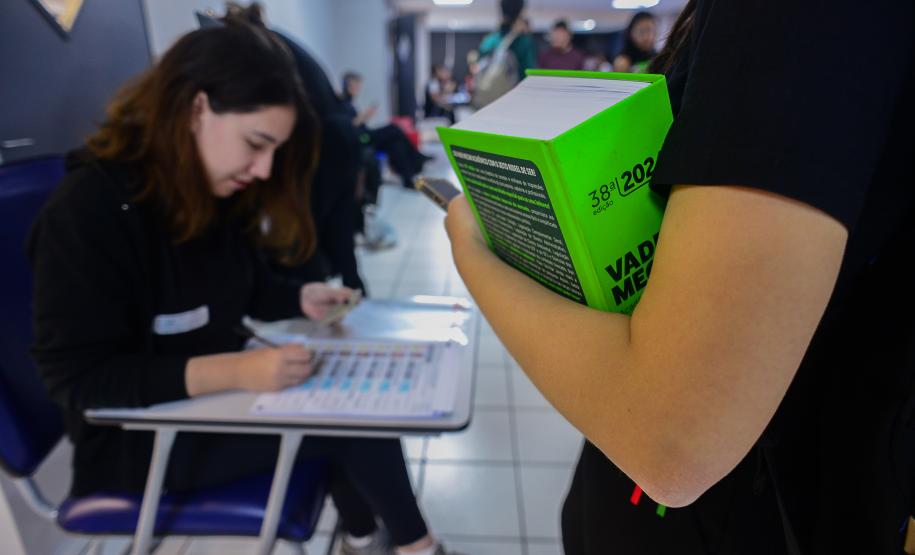 Imagem mostra braço de candidata segurando livro em close no primeiro plano enquanto fiscal confere seus documentos para ingressar na sala para do concurso.