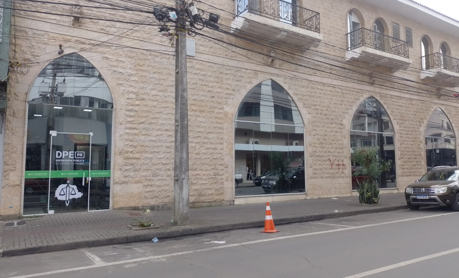 Imagem que mostra o prédio da nova sede da DPE em União da Vitória. O prédio possui fachada do andar térreo revestida em pedras de cor clara e marcada por grandes janelas e uma porta em formato de arco ogival. O andar superior também apresenta janelas em arco com sacadas e grades de ferro. À esquerda, uma porta de vidro exibe o logotipo e o nome da instituição. Através do vidro, é possível ver parte do interior da recepção.