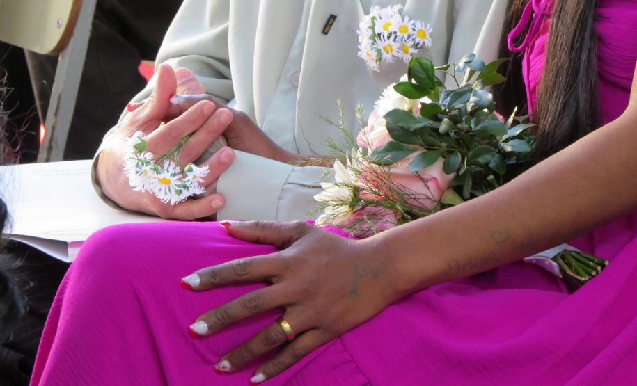 Uma imagem focada nas mãos de um casal. A noiva, usando um vestido vibrante de cor magenta, tem uma aliança dourada no dedo e as unhas pintadas. Sua mão repousa sobre a perna, próxima a um buquê. As mãos do noivo, que veste uma camisa clara, estão unidas e adornadas com um pequeno arranjo de margaridinhas amarrado em seu pulso.