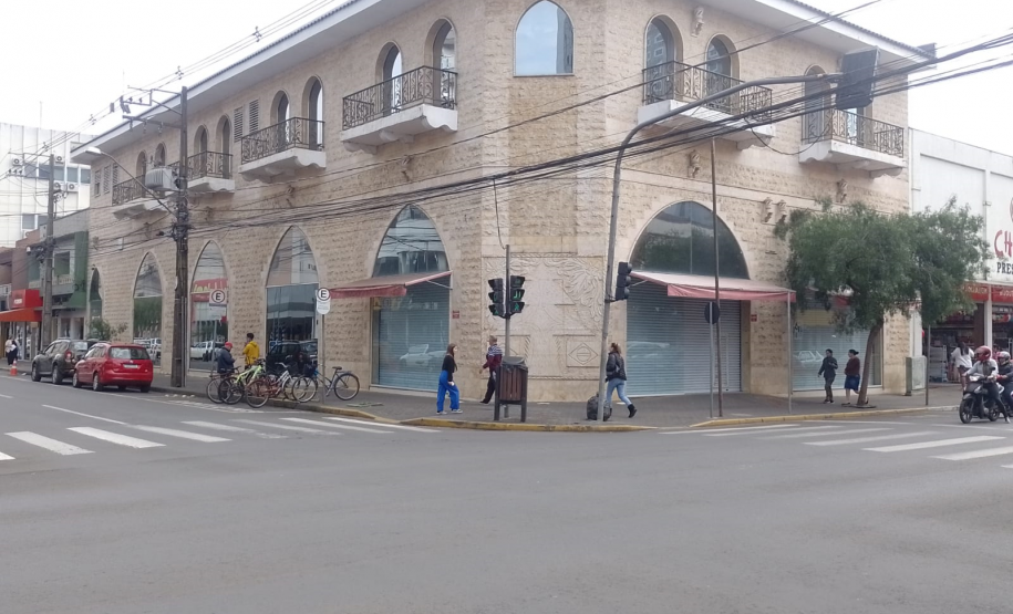 Imagem que mostra o prédio da nova sede da DPE em União da Vitória visto a partir de uma esquina. Há semáforos para carros e pedestres, e várias pessoas estão caminhando e atravessando a rua. Um grupo de bicicletas está estacionado na calçada. Carros estão parados na rua à esquerda e uma motocicleta passa pela direita. O céu está encoberto, indicando um dia nublado.