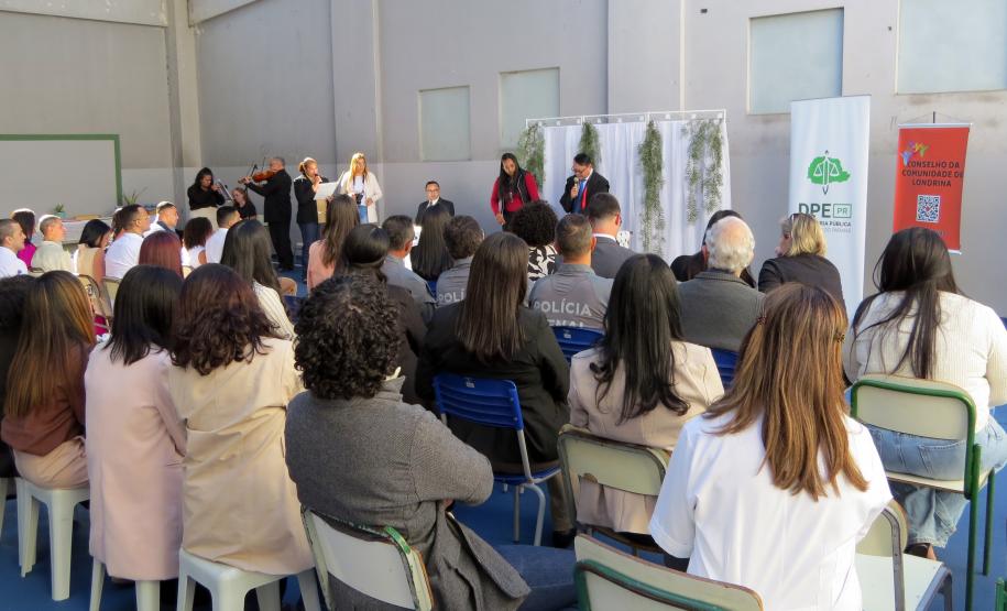 Esta foto oferece uma visão mais ampla da cerimônia, tirada de trás dos convidados. A plateia está sentada em cadeiras simples, observando os casais e o celebrante no palco. No lado direito, são visíveis dois banners: um do "Conselho da Comunidade de Londrina" e outro da Defensoria Pública do Estado do Paraná.