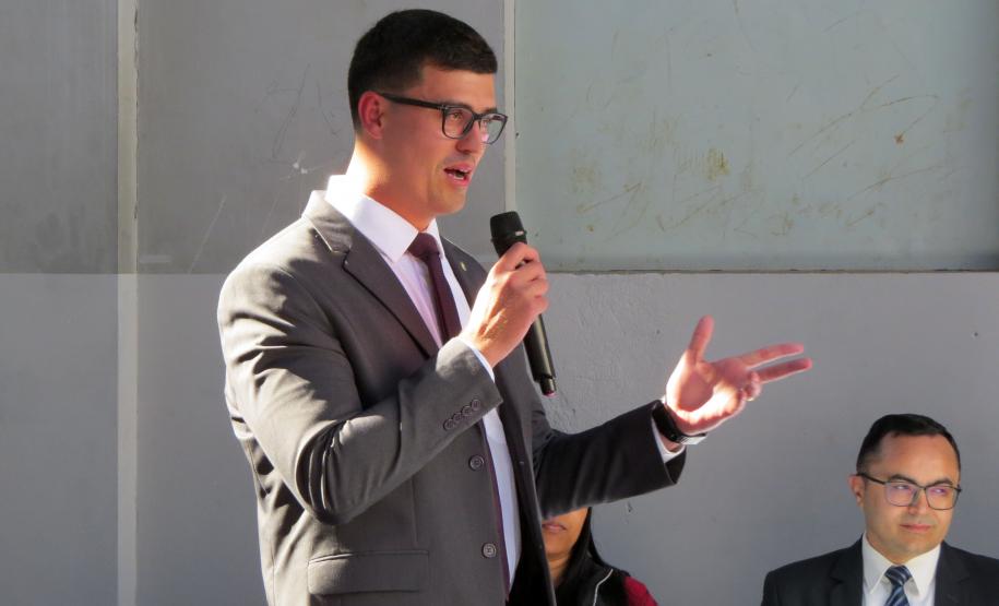 Imagem do defensor Pedro Bruzzi discursando com um microfone na mão. Ele veste um terno cinza e óculos, sua expressão e gestos indicam que ele está fazendo um pronunciamento como uma das autoridades do evento. Ao fundo, outra pessoa pode ser vista sentada.