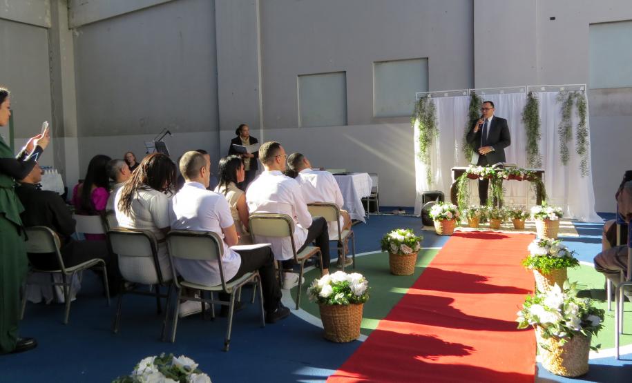 A imagem mostra a cerimônia de casamento em andamento. Vários casais estão sentados na primeira fila, com os homens vestindo camisas brancas. Ao fundo, convidados assistem. O pastor, de terno, fala ao microfone em um pequeno palco decorado com cortinas brancas e folhagens. Um tapete vermelho demarca o corredor central, ladeado por cestos com flores brancas. A luz do sol forte cria sombras pronunciadas.