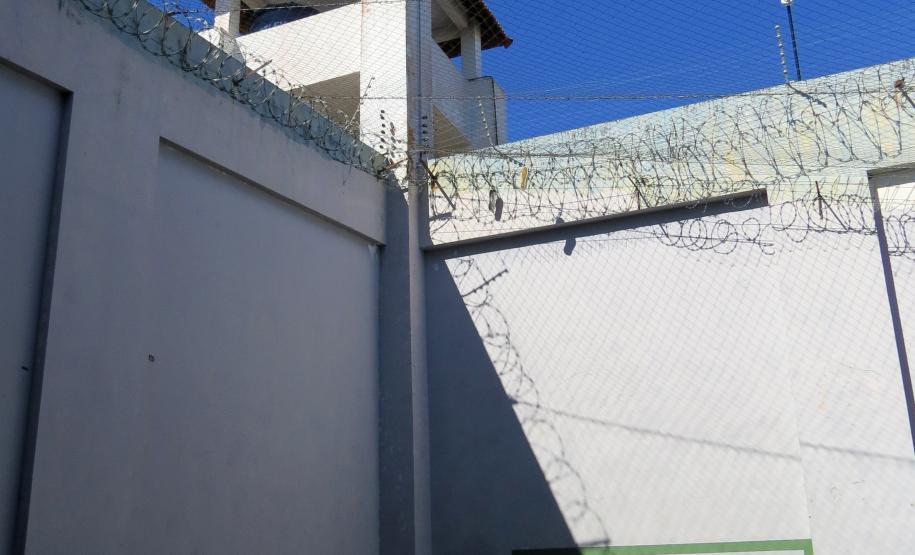 Imagem que captura o cenário da cerimônia. No pátio da prisão, com o chão pintado de azul, erguem-se altos muros de concreto cinza, coroados com arame farpado e uma rede de proteção que se estende sob o céu azul. Ao fundo, uma guarita de vigilância se destaca. Em um canto do pátio, um pequeno altar foi montado com um arco circular dourado, decorado com flores brancas e um tecido esvoaçante, contrastando a delicadeza da celebração com a severidade do ambiente.