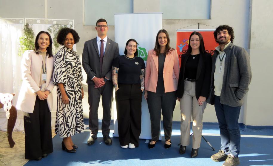 Um grupo de sete pessoas posa para uma foto. São defensores e assessores da DPE-PR. Eles estão no pátio da prisão, em frente banners da Defensoria e do Conselho da Comunidade. As vestimentas são formais e a atmosfera é de satisfação e dever cumprido.