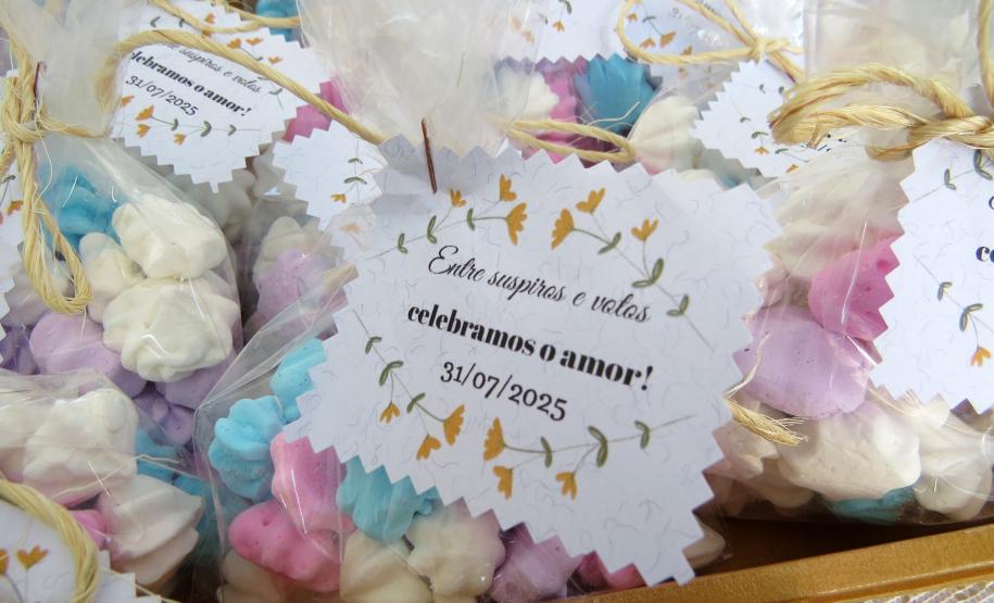 Esta é uma foto das lembrancinhas do casamento. Vários saquinhos transparentes estão cheios de suspiros coloridos em tons de rosa, azul, lilás e branco. Cada saquinho é amarrado com um laço de fio de ráfia e possui uma etiqueta de papel branco com bordas onduladas. A etiqueta, decorada com ramos de flores amarelas, traz a mensagem: "Entre suspiros e votos celebramos o amor! 31/07/2025".