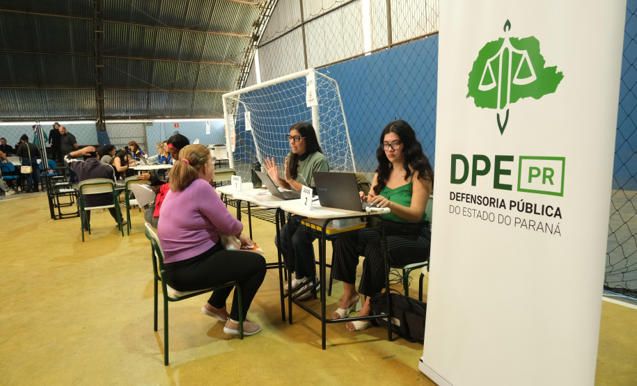Imagem que mostra um ginásio de esportes adaptado para o atendimento. Uma mulher de blusa roxa é acolhida por duas integrantes da equipe da Defensoria Pública do Paraná. Enquanto uma das atendentes dialoga e gesticula, a outra opera um notebook. Há um banner da DPE-PR, em primeiro plano à direita.