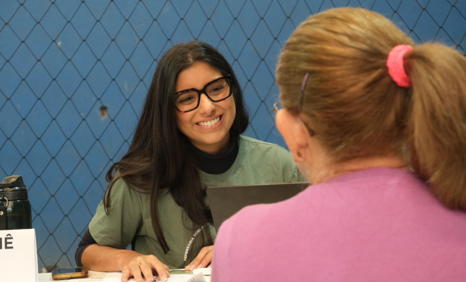 Imagem focada no rosto de uma das novas defensoras públicas que está sentada a uma mesa, vista de frente. Ela é uma mulher de pele clara, com cabelos pretos, longos e lisos, e usa óculos de armação preta e grossa. Ela olha para a direita, na direção de uma outra pessoa, com um sorriso largo e acolhedor. Ela atende uma mulher que tem cabelos loiros presos em um rabo de cavalo com um elástico rosa e veste uma blusa de cor magenta.