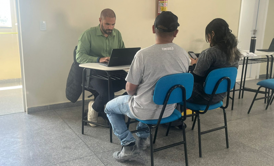 Imagem que mostra uma cena de atendimento individual. Três pessoas estão agrupadas em torno de uma pequena mesa branca. À esquerda, de frente para a mesa, está o atendente. Ele é um homem de pele morena, com cabelo escuro bem curto e barba. Veste uma camisa social de manga longa e está concentrado. Do outro lado da mesa, de costas para a câmera, estão duas pessoas sentadas em cadeiras com assentos e encostos azuis, um homem e uma mulher.