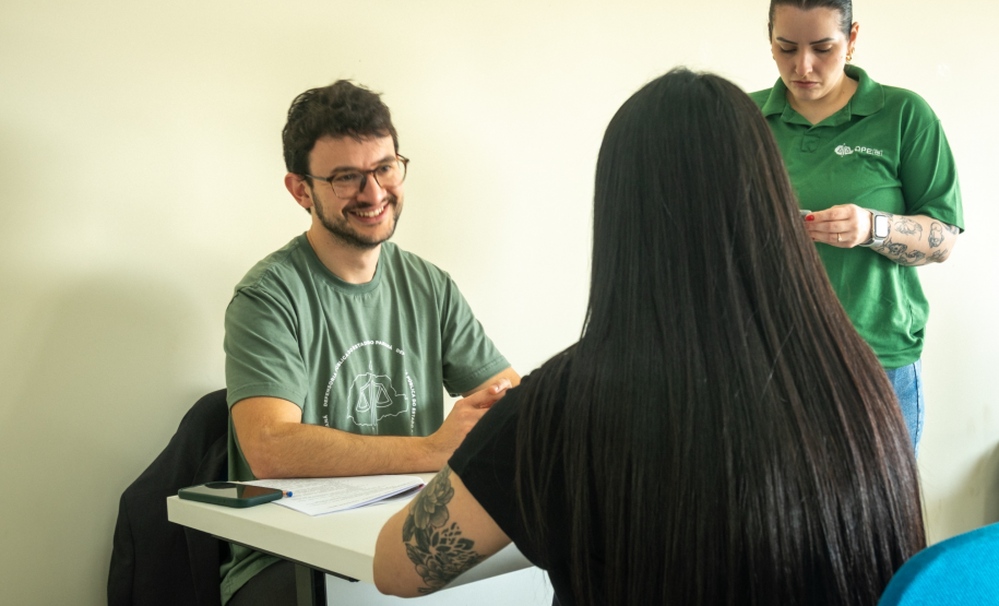 Imagem que mostra um atendimento com três pessoas em uma sala de paredes claras. Em foco, sentado a uma mesa branca, está um dos novos defensores públicos, um homem jovem, de pele clara, com cabelo escuro e encaracolado, barba e óculos. Ele sorri de forma aberta e gentil para a pessoa que atende: uma mulher de cabelos longos, lisos e pretos. Em pé, ao lado direito e um pouco atrás do defensor, uma segunda integrante da equipe, de cabelo preso e vestindo uma camisa polo verde, observa o atendimento.