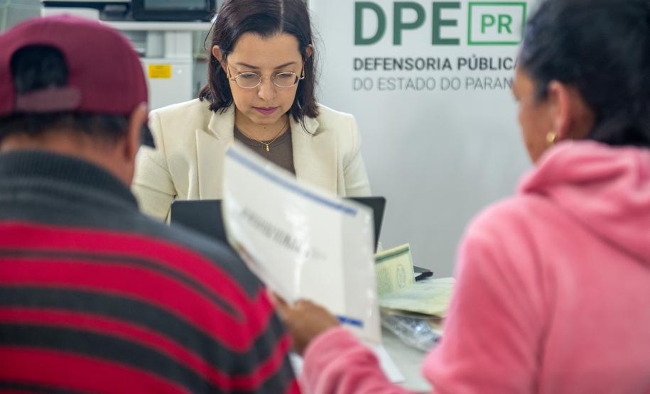 Assistidos aparecem de costas desfocados entregando documentos à servidora da DPE-PR em Mutirãop de Conciliação.