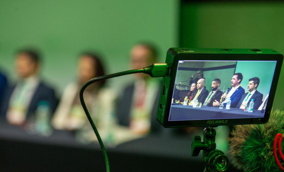 Imagem mostra tela da filmadora em primeiro plano e ao fundo a mesa de abertura do VI Conajuri em Curitiba,