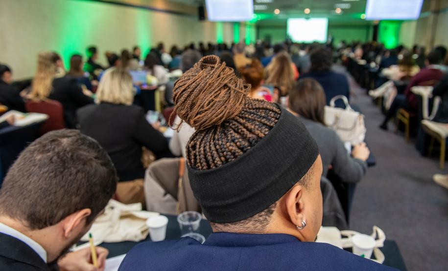 Homem negro com tranças e homem branco tomando notas em caderno, ambos de costas em primeiro plano, e ao fundo se vê a sala lotada de defensores de too o país na abertura di Conajuri.
