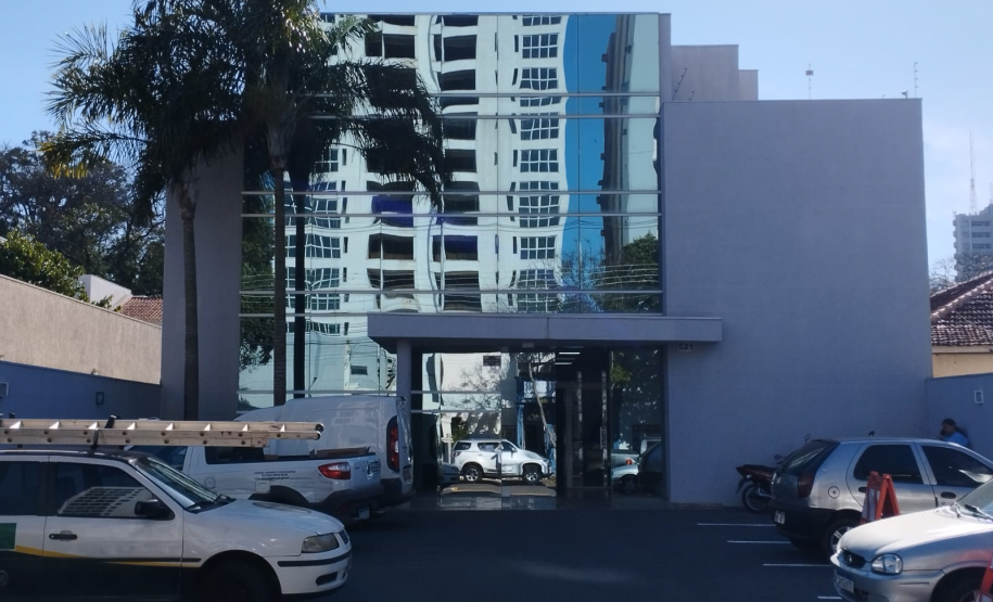 Imagem que mostra a fachada da sede da Defensoria Pública em Cianorte, em um dia claro e ensolarado. O prédio é uma construção moderna de dois andares, com uma grande seção central de vidro espelhado. As laterais do prédio são de alvenaria, pintadas em um tom de cinza-claro. Na frente do edifício há um estacionamento com alguns carros parados. À esquerda do prédio, uma palmeira alta se destaca contra o céu.