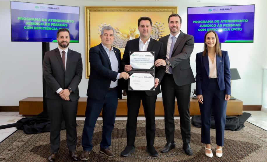 Um grupo de cinco pessoas, quatro homens e uma mulher, posa para uma foto em uma sala formal. Os dois homens no centro, o governador e o DPG, seguram e exibem dois certificados emoldurados para a câmera.