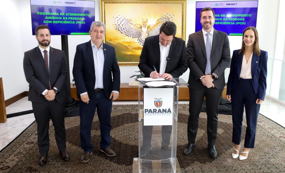 Em uma sala formal, quatro pessoas em pé observam o governador no centro, que se inclina sobre um púlpito transparente para assinar um documento.