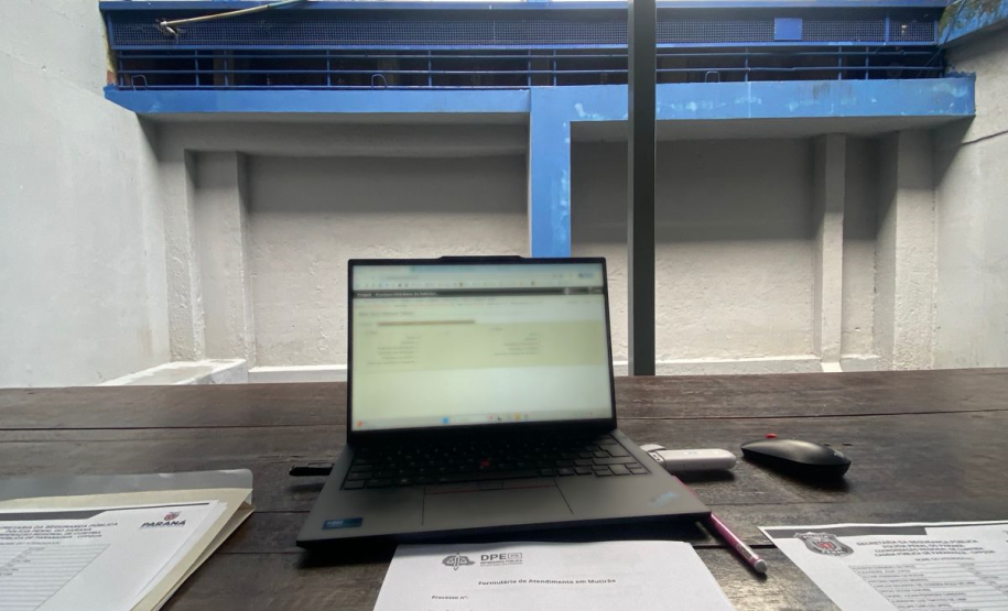 A imagem é uma visão em primeiro plano de uma mesa de trabalho de madeira. Sobre a mesa, há um laptop aberto e formulários de questionário para entrevistas. O ambiente é o pátio interno da cadeia, com paredes de concreto e estruturas pintadas de azul na parte superior.