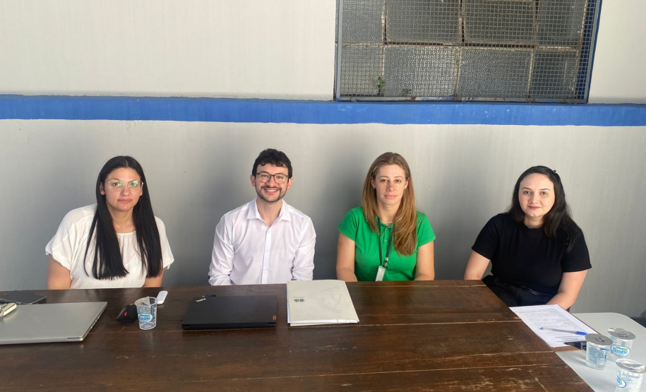Quatro pessoas, três mulheres e um homem, posam para uma foto, sentados lado a lado atrás de uma mesa de madeira. Eles estão em um local com parede branca e uma faixa azul, com uma janela gradeada ao fundo.