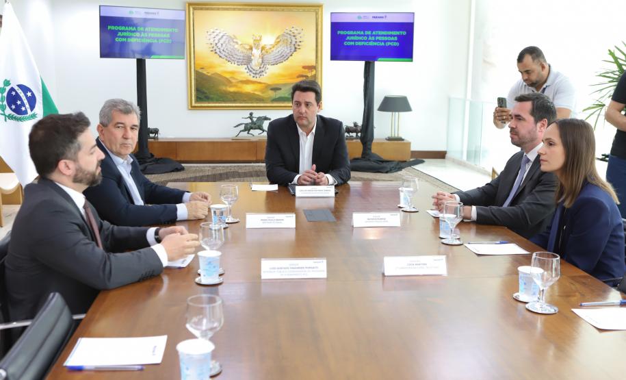 Cinco pessoas estão sentadas em uma mesa de reunião de madeira em uma sala formal. Ao fundo da sala, há duas telas de televisão ligadas com o título de um programa e, à esquerda, a bandeira do Paraná em um mastro. Um homem que está à direita aponta um celular para a cena.