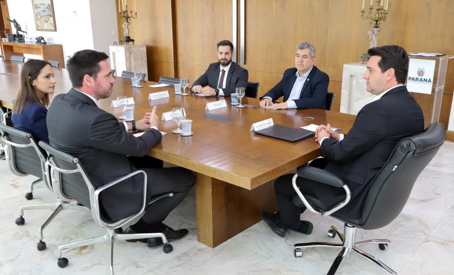 Cinco pessoas em trajes sociais, sendo quatro homens e uma mulher, estão sentadas ao redor de uma grande mesa de madeira em uma sala de reuniões. Elas estão em uma conversa formal, com copos de água e papéis sobre a mesa.