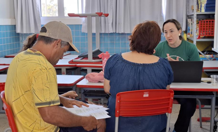 Dentro de uma sala de aula, um casal é atendido por uma servidora da Defensoria Pública de camisa verde. O homem, de camiseta amarela e boné, lê alguns papéis, enquanto a mulher, de blusa jeans e de costas para a foto, ouve a explicação da servidora.