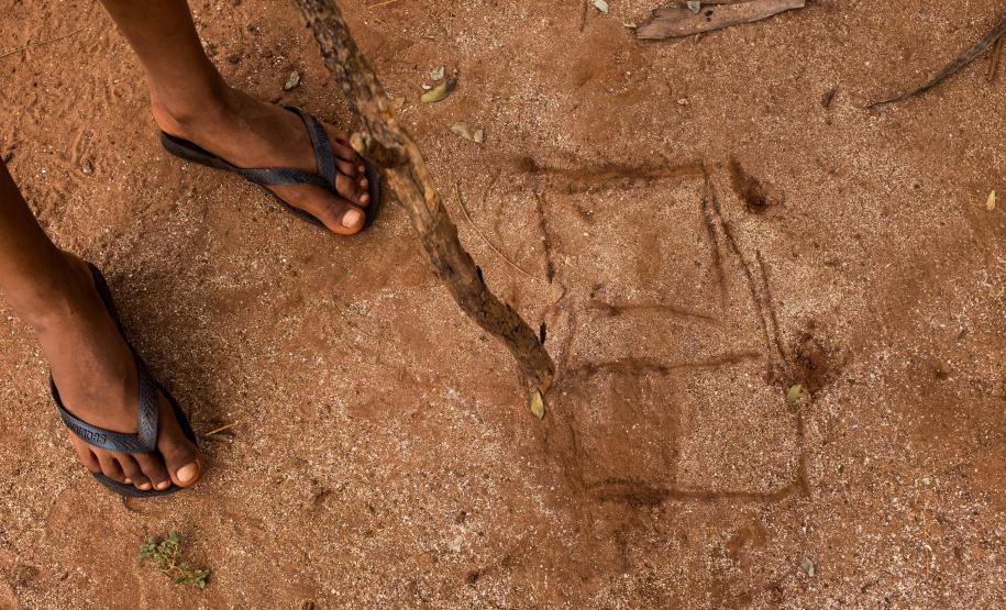 Imagem mostra detalhe dos pés de uma criança de chinelo usando um galho de madeira para desenhar um cubo na terra avermelhada.