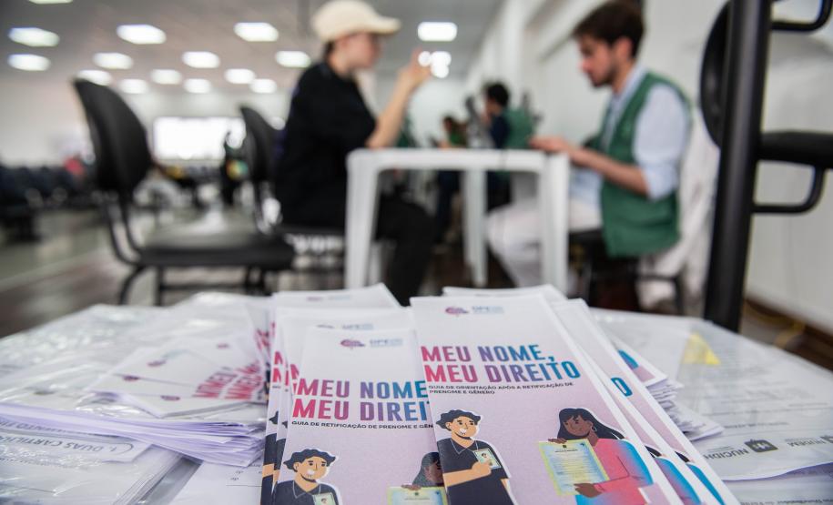 Imagem mostra panfletos do Meu Nome Meu Direito em primeiro plano e ao fundo aparece um assistido recebendo atendimento. Em agosto o projeto chegou aos mil atendimentos em todo o Paraná.