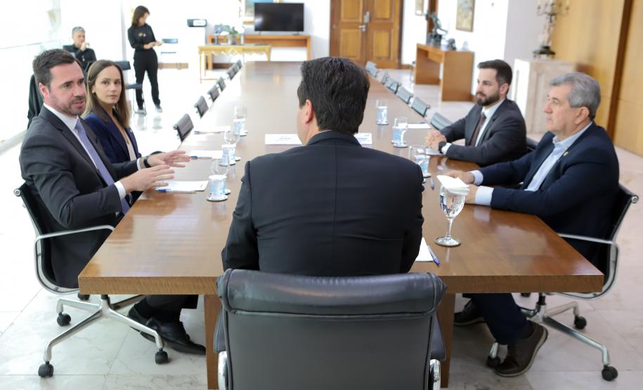 A foto é tirada por trás do governador, sentado na cabeceira de uma longa mesa de reuniões de madeira. De cada lado da mesa, duas pessoas em trajes sociais participam da conversa, sendo que um homem à esquerda gesticula enquanto fala. Ao fundo da sala ampla e formal, outras duas pessoas estão em pé.