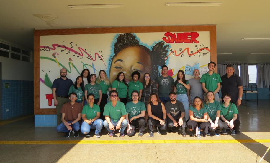 Uma foto de grupo com cerca de vinte pessoas, a equipe da Defensoria Pública, do CRAS Rural e da Escola Municipal posando em um pátio coberto de uma escola. Eles estão em pé e agachados em frente a um grande mural colorido pintado na parede ao fundo.