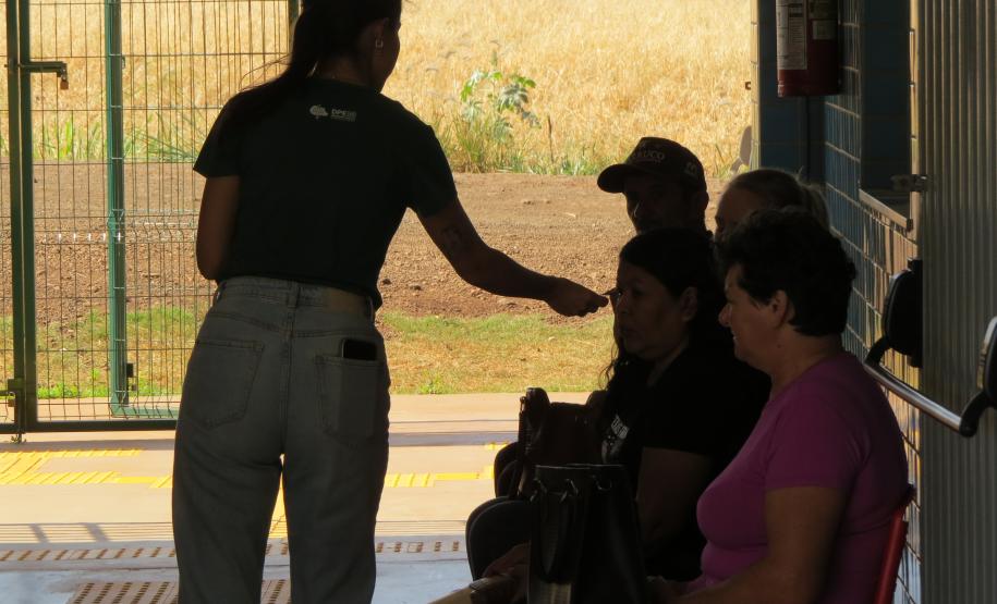 Vista de dentro de um pátio, uma servidora está em pé, conversando com um grupo de três pessoas sentadas em um banco. Ao fundo, através de uma grade, vê-se um campo aberto com vegetação seca sob a luz do sol.
