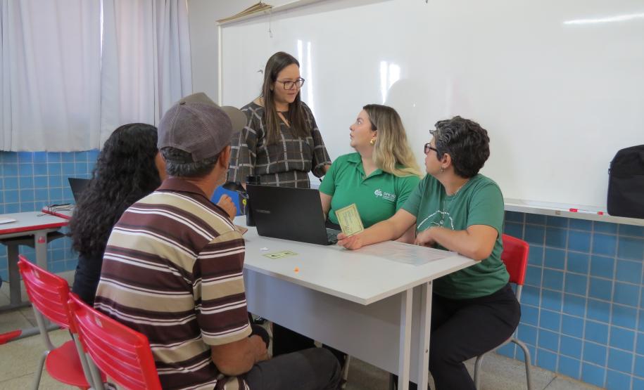 Em uma sala de aula, cinco pessoas estão reunidas ao redor de uma mesa branca com um laptop. Duas servidoras em camisas verdes e uma terceira mulher em pé atendem a um casal, que está sentado de costas para a câmera, em cadeiras vermelhas.