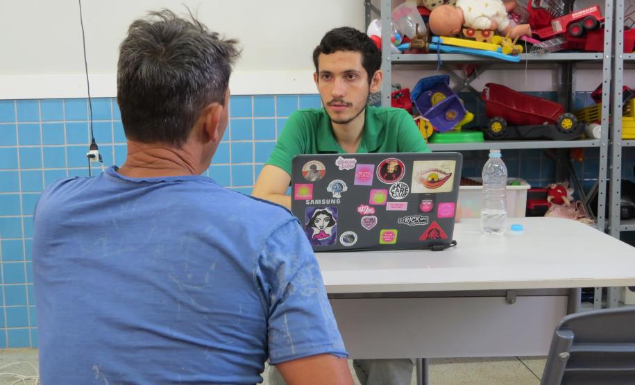 Dois homens conversam em uma mesa, um de frente para o outro. O homem de costas, com uma camisa azul, fala com um servidor mais jovem de camisa verde. Entre eles, sobre a mesa, há um laptop com vários adesivos. Ao fundo, prateleiras com brinquedos.