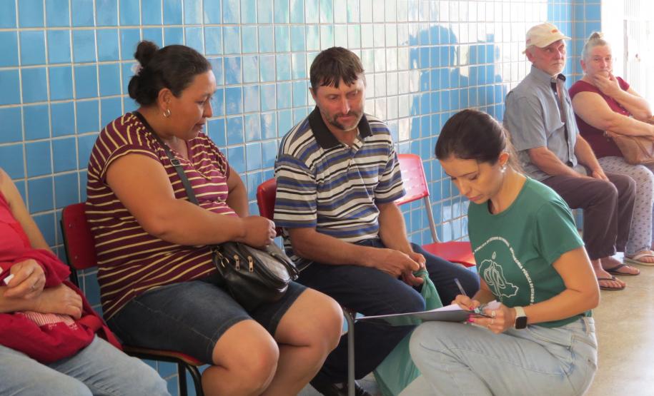 Em um corredor com parede de azulejos azuis, pessoas aguardam sentadas. Em primeiro plano, uma servidora de camisa verde está agachada, anotando informações em uma prancheta enquanto conversa com um casal sentado em cadeiras vermelhas.