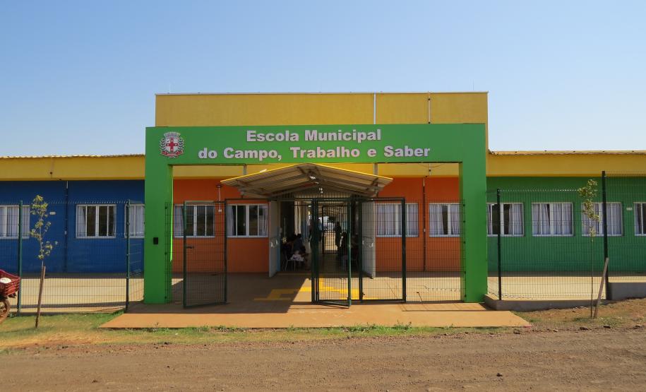 A foto mostra a fachada colorida de uma escola rural, com paredes pintadas em azul, laranja e verde. Um grande portal verde sobre a entrada tem a inscrição "Escola Municipal do Campo, Trabalho e Saber". O dia está ensolarado, com céu azul e sem nuvens.