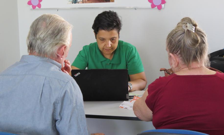 Um casal de idosos, vistos de costas, está sentado em frente a uma mesa de atendimento. Eles são atendidos por uma servidora de camisa verde, que está sentada do outro lado da mesa, concentrada na tela de um laptop.