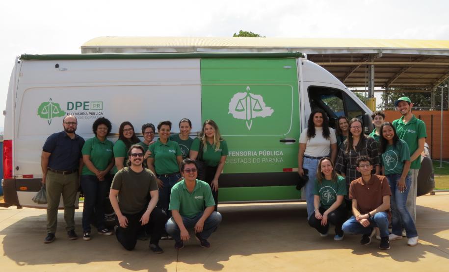 Um grupo grande de pessoas, a equipe da Defensoria Pública, posa para uma foto em frente a uma van branca. O veículo tem uma faixa verde e o logotipo da DPE PR e está estacionado no estacionamento da escola.
