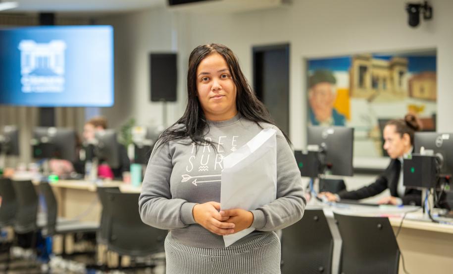 Uma mulher jovem segura seus documentos e aparece no centro da imagem.