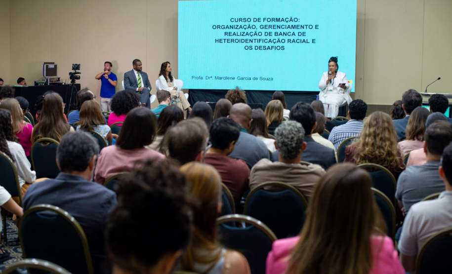 Um auditório cheio com a plateia sentada de costas para a foto, voltada para um palco. No palco, uma mulher de branco fala ao microfone ao lado de uma tela projetando o título de um curso sobre heteroidentificação racial.