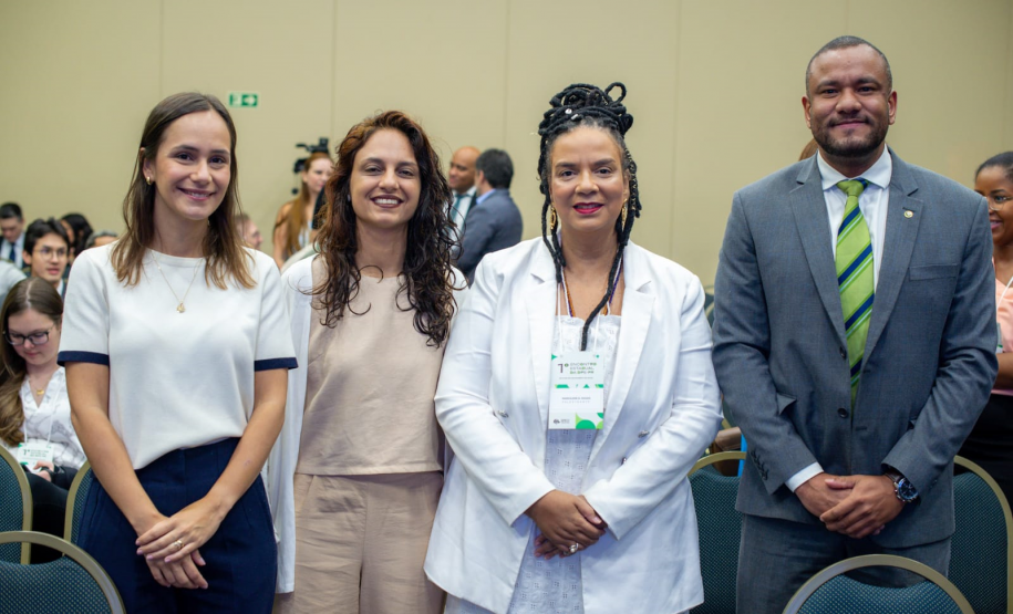 Quatro pessoas posam lado a lado para uma foto em um ambiente interno. Da esquerda para a direita: uma mulher de blusa branca, uma mulher de roupa bege, uma mulher negra com tranças e roupa branca, e um homem negro de terno cinza e gravata verde.