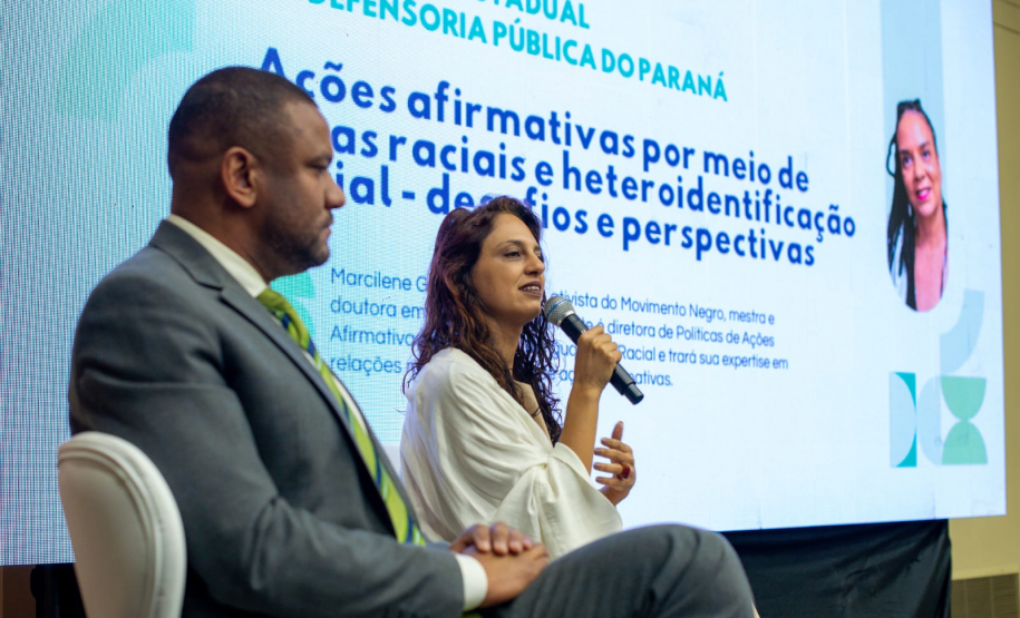 Uma mulher de cabelos longos e blusa branca fala ao microfone, sentada ao lado de um homem de terno. Atrás deles, um telão exibe o texto "Ações afirmativas por meio de cotas raciais e heteroidentificação".