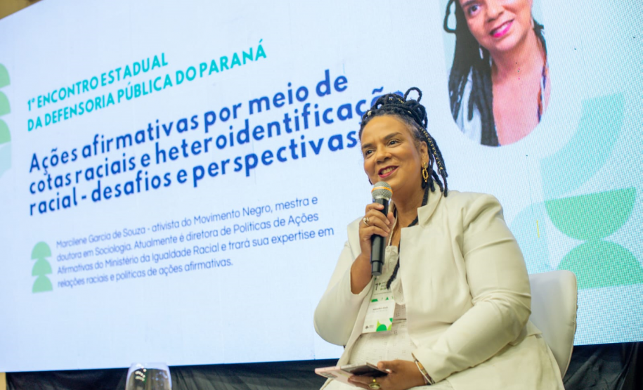Uma mulher negra com tranças e roupa branca sorri enquanto fala ao microfone, sentada no palco. Ao fundo, uma tela projeta sua foto, nome e o tema da palestra sobre ações afirmativas e cotas raciais.